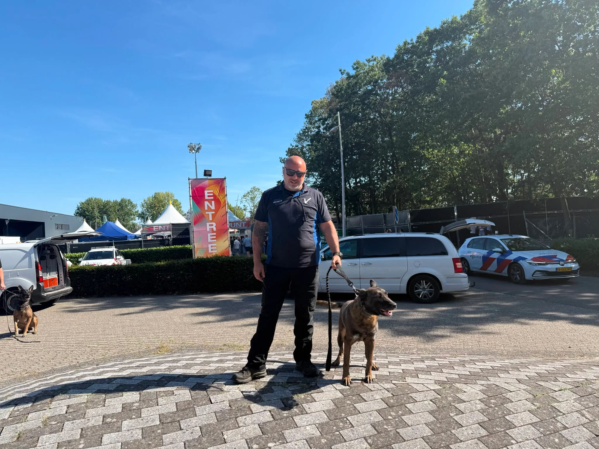 Hondengeleider van SSG met een Mechelse herder aan de lijn bij de entree van een evenemententerrein